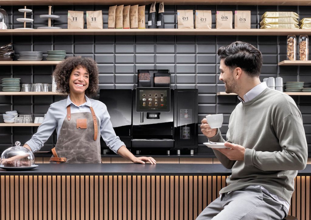 Franke Coffee Systems, man sitting at the counter of a coffee shop drinking coffee served by woman shop assistant, fully automatic coffee machine Franke 