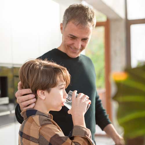 Young boy drinking filtered water from a glass with father standing next to him with his hand on back of head.